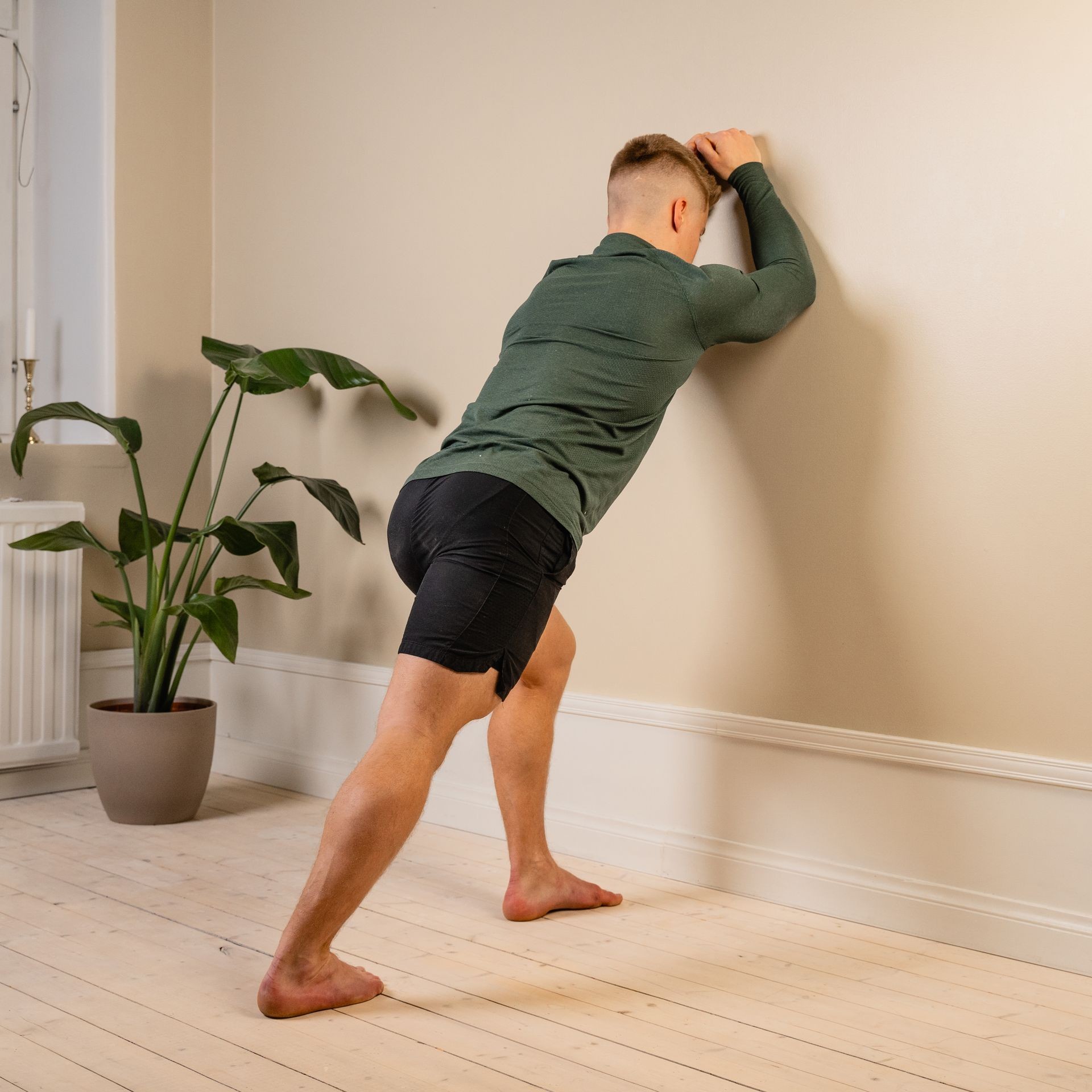 Person performing a calf stretch against a wall in a room with wooden floor and a plant nearby.