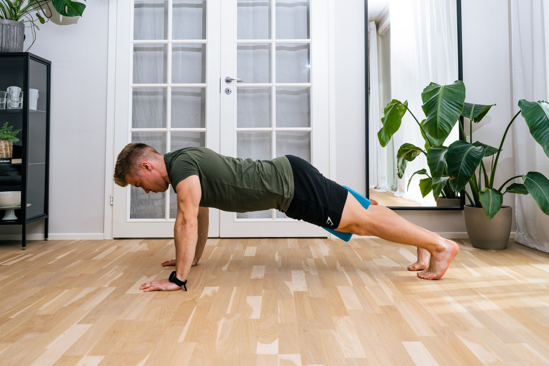 Person performing a push-up on a wooden floor in a bright room with plants.