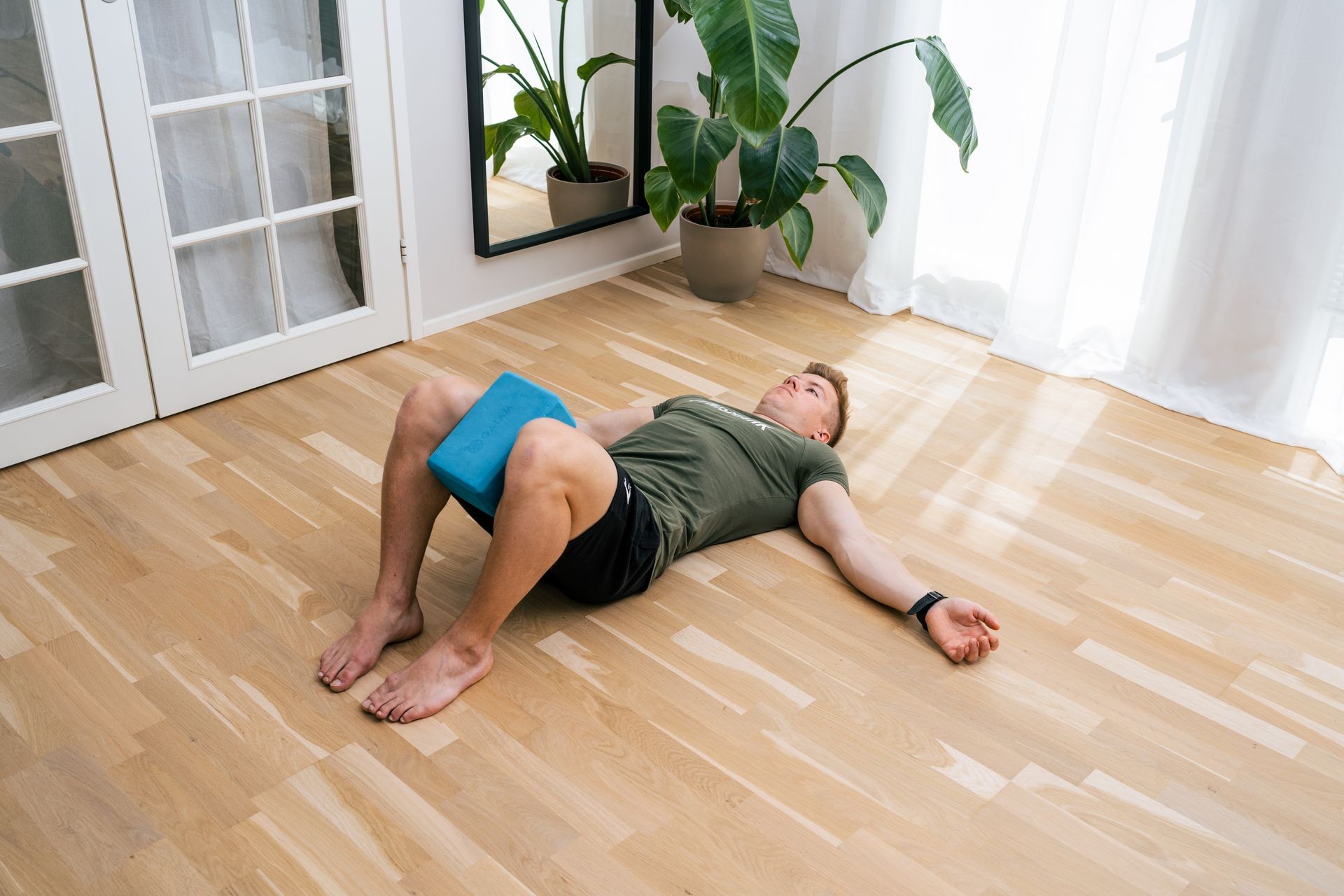 Person lying on the floor with a yoga block between knees in a sunlit room with plants.