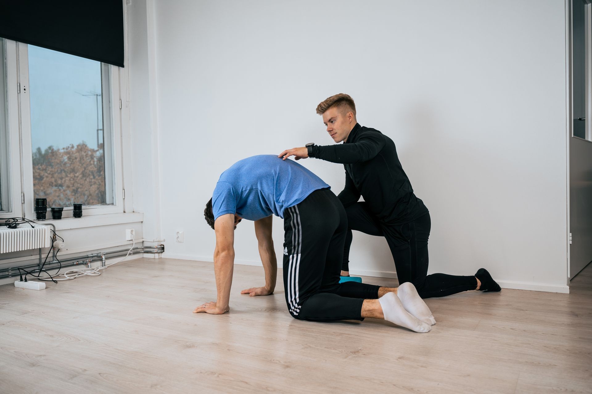 A physical therapist assisting a man in performing a stretching exercise on the floor in a minimalist room.