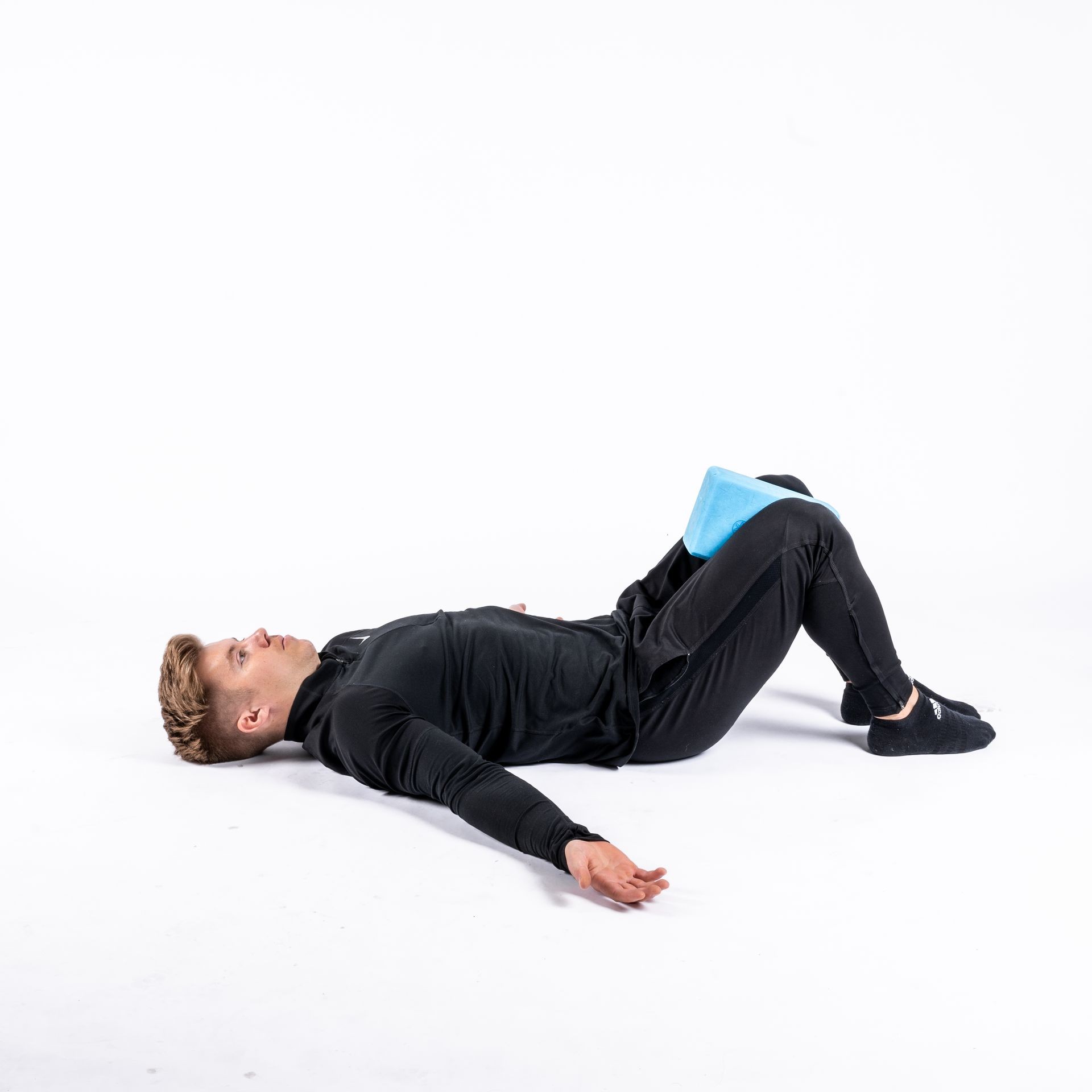 Person lying on back with knees bent and foam block between knees, exercising on white background.