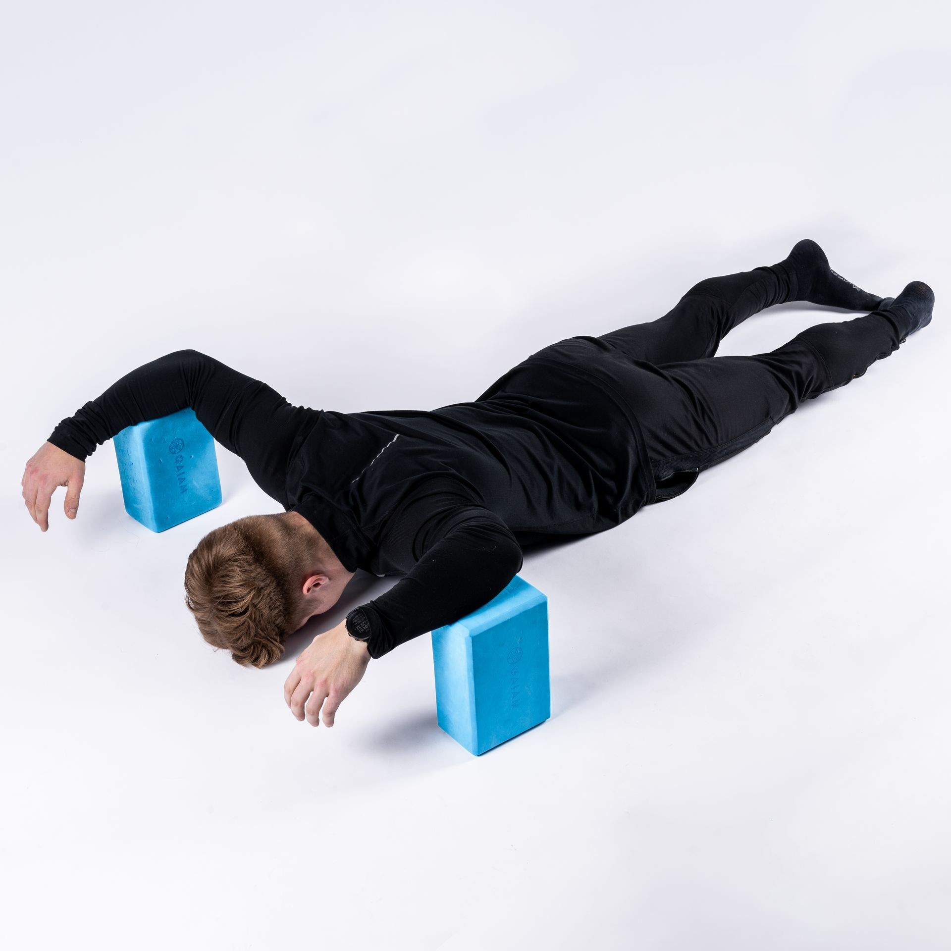 Person in black workout attire lying face down with arms raised, supported by two blue foam blocks.